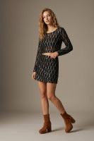 A woman modeling a long-sleeve black mini dress with a textured geometric pattern, paired with brown ankle boots, against a solid warm beige studio background.
